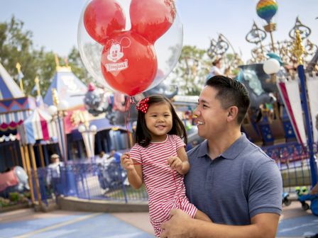 A man holding a young child with a Mickey Mouse balloon at an amusement park, surrounded by colorful attractions and decorations, both are smiling.