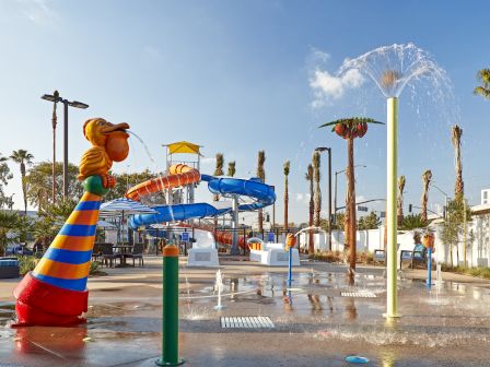 A colorful water park with slides, sprinklers, and palm trees under a clear sky can be seen.