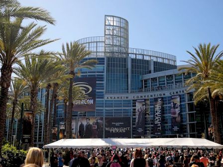 The image shows a large crowd in front of the Anaheim Convention Center with palm trees and banners advertising a convention.