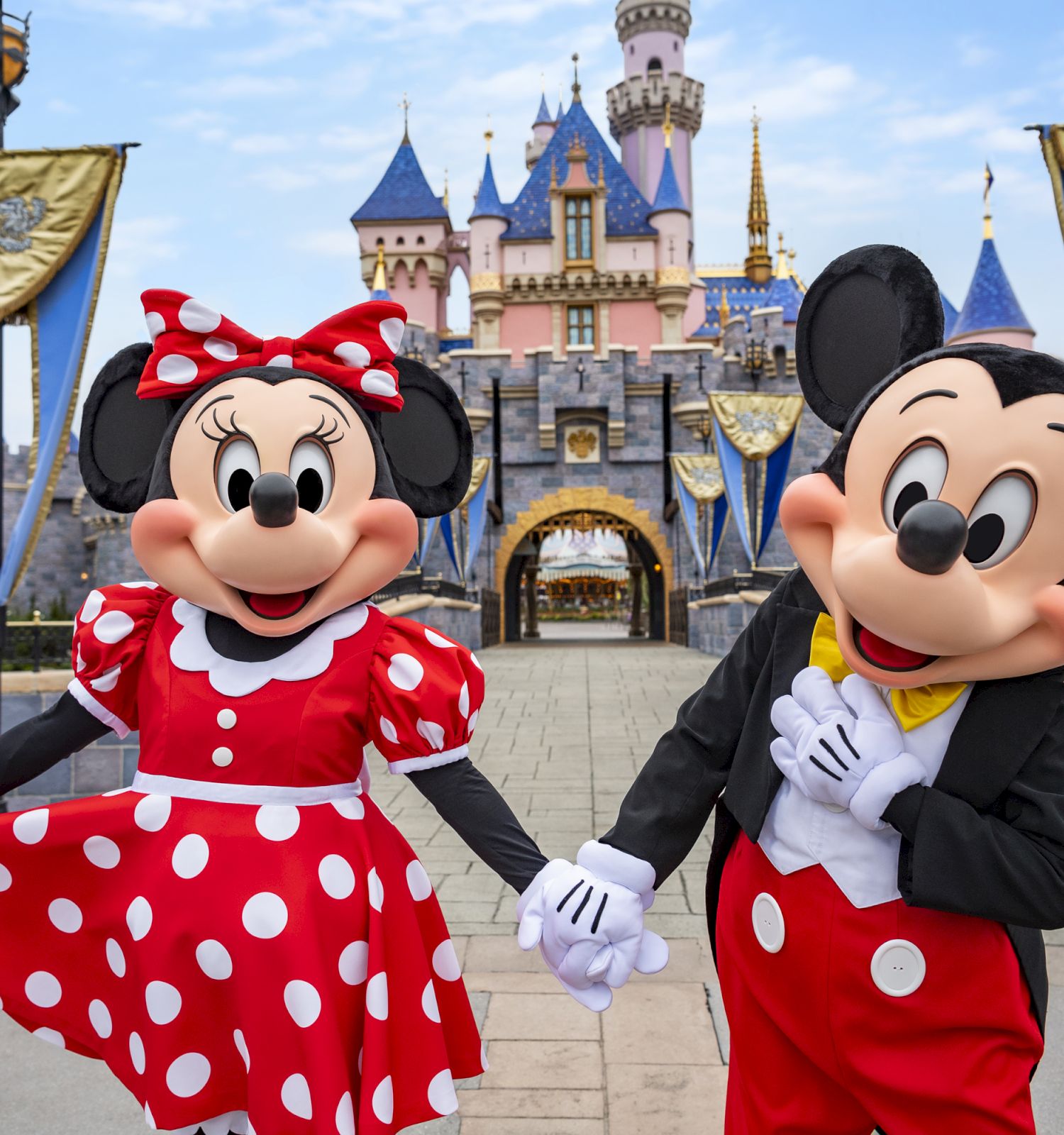 Two iconic animated characters, one in red polka dot dress, the other in red shorts, holding hands in front of a castle with blue and gold banners.