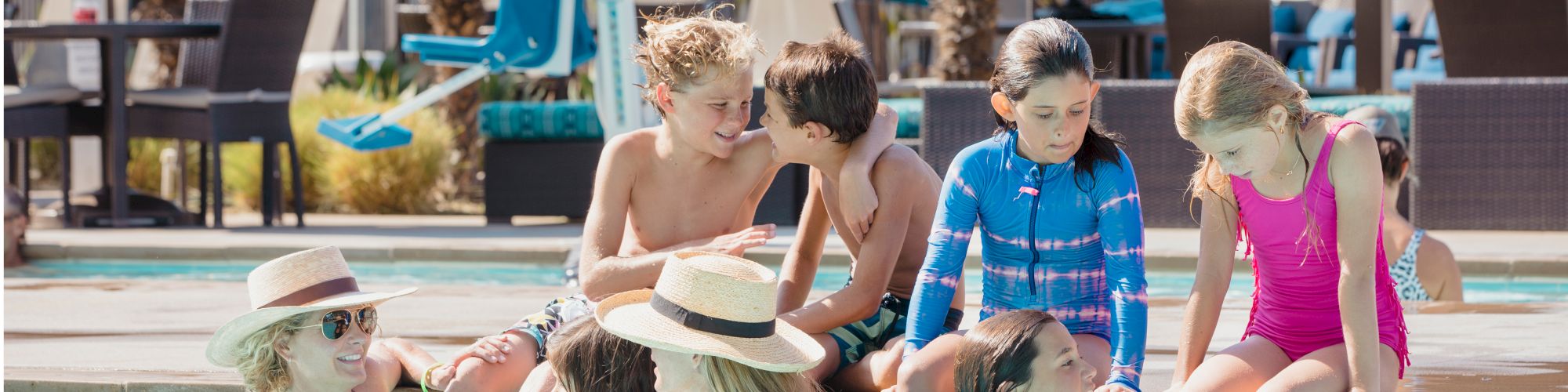 A group of people, including children and adults, are enjoying their time together at a poolside with some in the pool and others sitting at the edge.