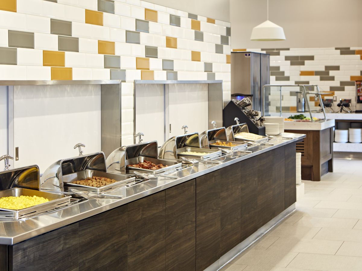 The image shows a buffet setup with several food trays, a modern tiled wall, and a section with utensils and dishes in the background.