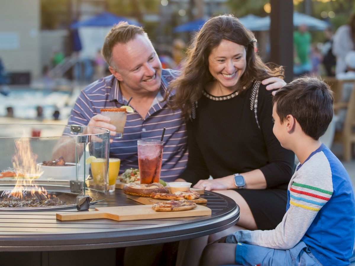 A family is enjoying an outdoor meal at a restaurant, sitting around a round table with drinks and food, with evening light in the background.