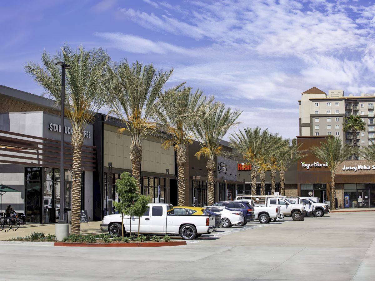 A commercial plaza with palm trees in front, featuring stores like Starbucks and Yogurtland, with trucks and cars parked in the lot.