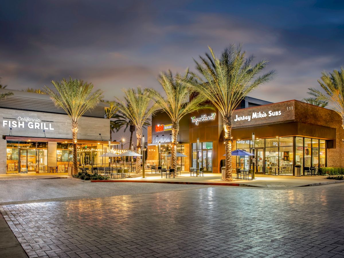 The image shows an outdoor shopping area at dusk, with restaurants like Fish Grill and Jersey Mike's Subs, and palm trees lining the walkway.