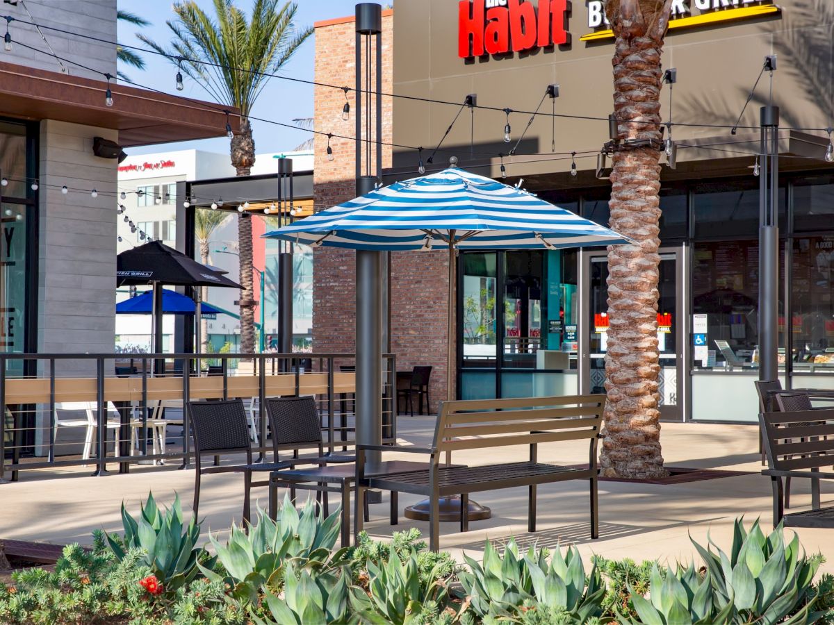 An outdoor seating area with benches, tables, and a blue-striped umbrella is in front of a restaurant, surrounded by plants and palm trees.