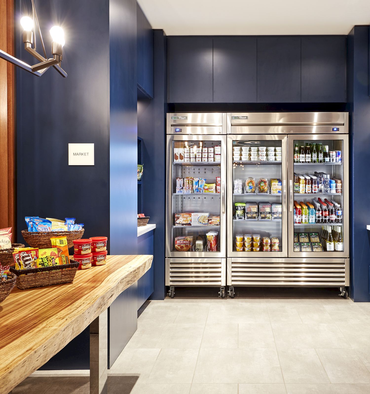 A small store featuring a wooden table with snack baskets, shelves with various items, and stainless steel refrigerators stocked with beverages and packaged goods.