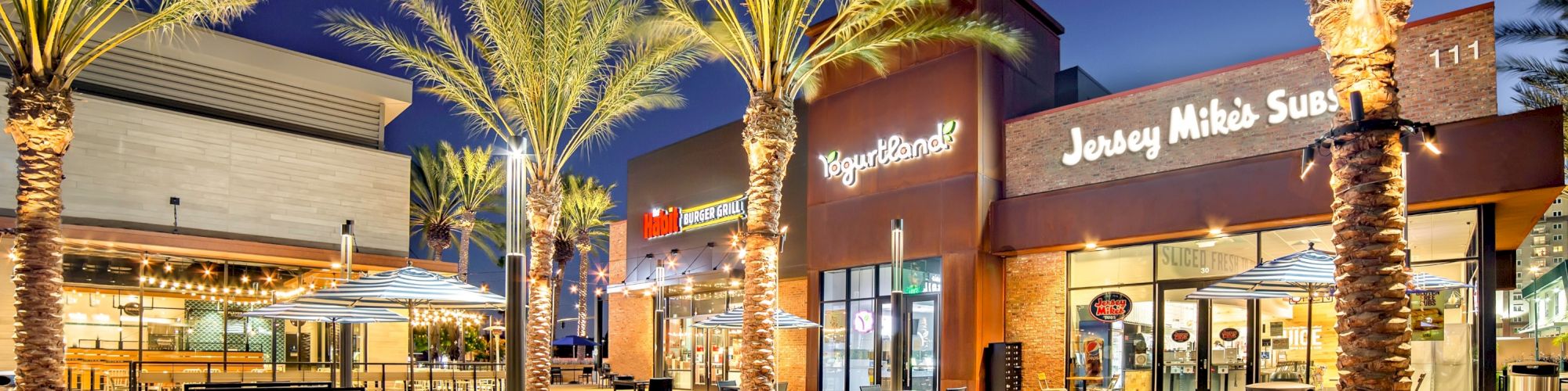 The image shows an outdoor shopping area at night with palm trees, restaurants with outdoor seating, and bright signage for eateries like Jersey Mike's Subs.