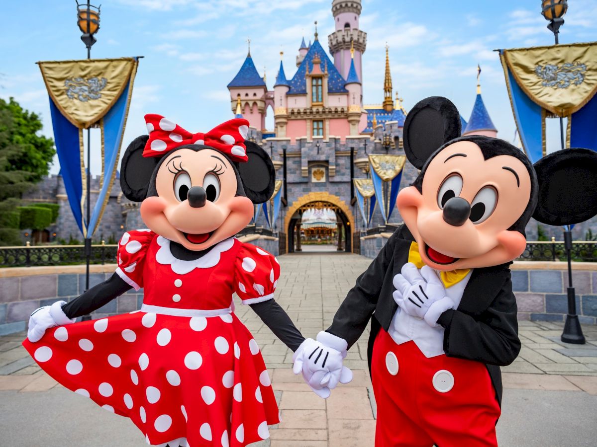 The image shows two characters in costume, one in a red polka dot dress and the other in red pants with white buttons, standing in front of a castle.