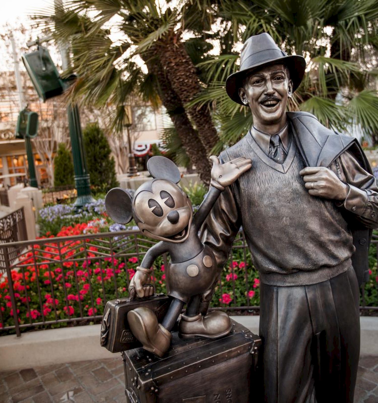 A statue of a man holding a hat next to Mickey Mouse, set in a colorful garden area with flowers and palm trees, is shown in the image.