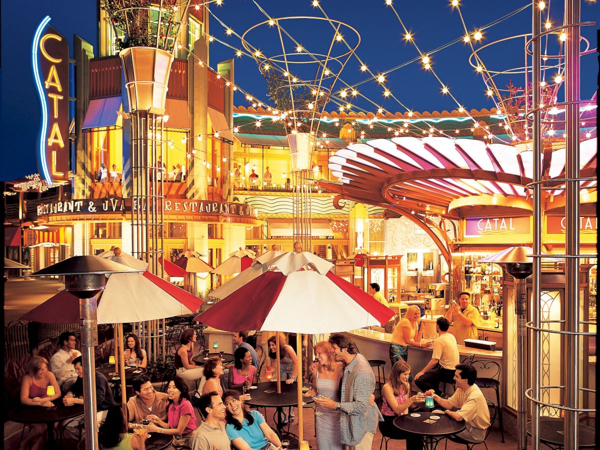 An outdoor dining area at night with people sitting under red and white umbrellas. The background features colorful lighting and structures.