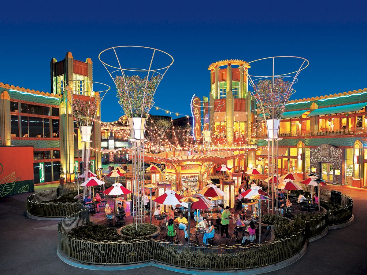 The image depicts a vibrant outdoor shopping and dining area with colorful lights and umbrellas, surrounded by Art Deco-style buildings.