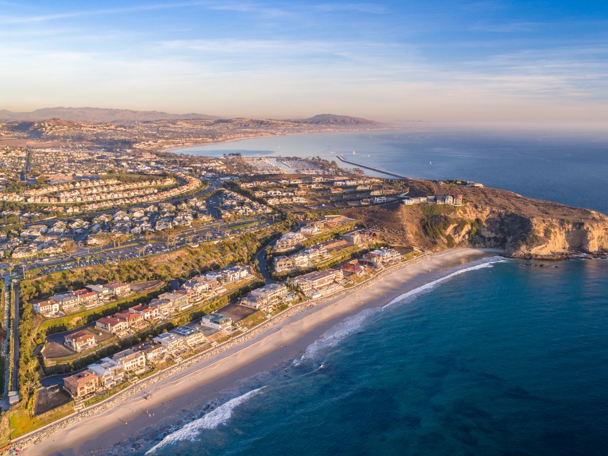 The image shows an aerial view of a coastal city with beachfront houses, a coastline, and a pier extending into the ocean, under a clear sky.