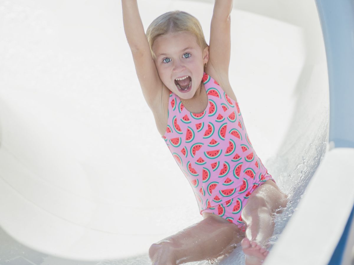 A child in a swimsuit is joyfully sliding down a water slide with arms raised and mouth open, appearing excited.