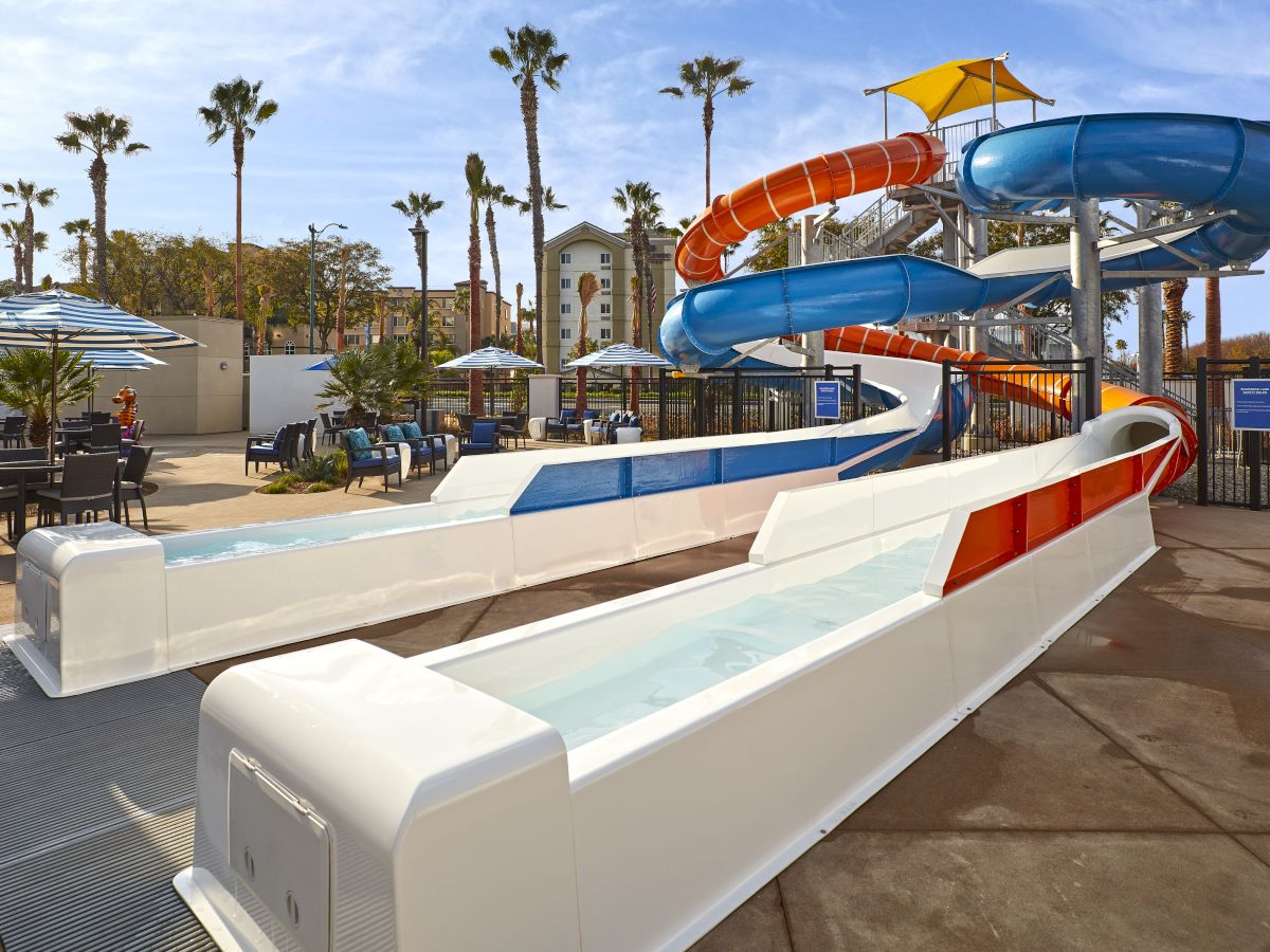 The image shows a pair of colorful water slides with a pool, situated in an outdoor area with palm trees and umbrellas.