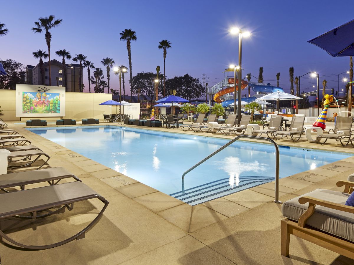 A pool area is shown with lounge chairs, umbrellas, and a screen displaying a movie. The setting is illuminated, suggesting evening time.