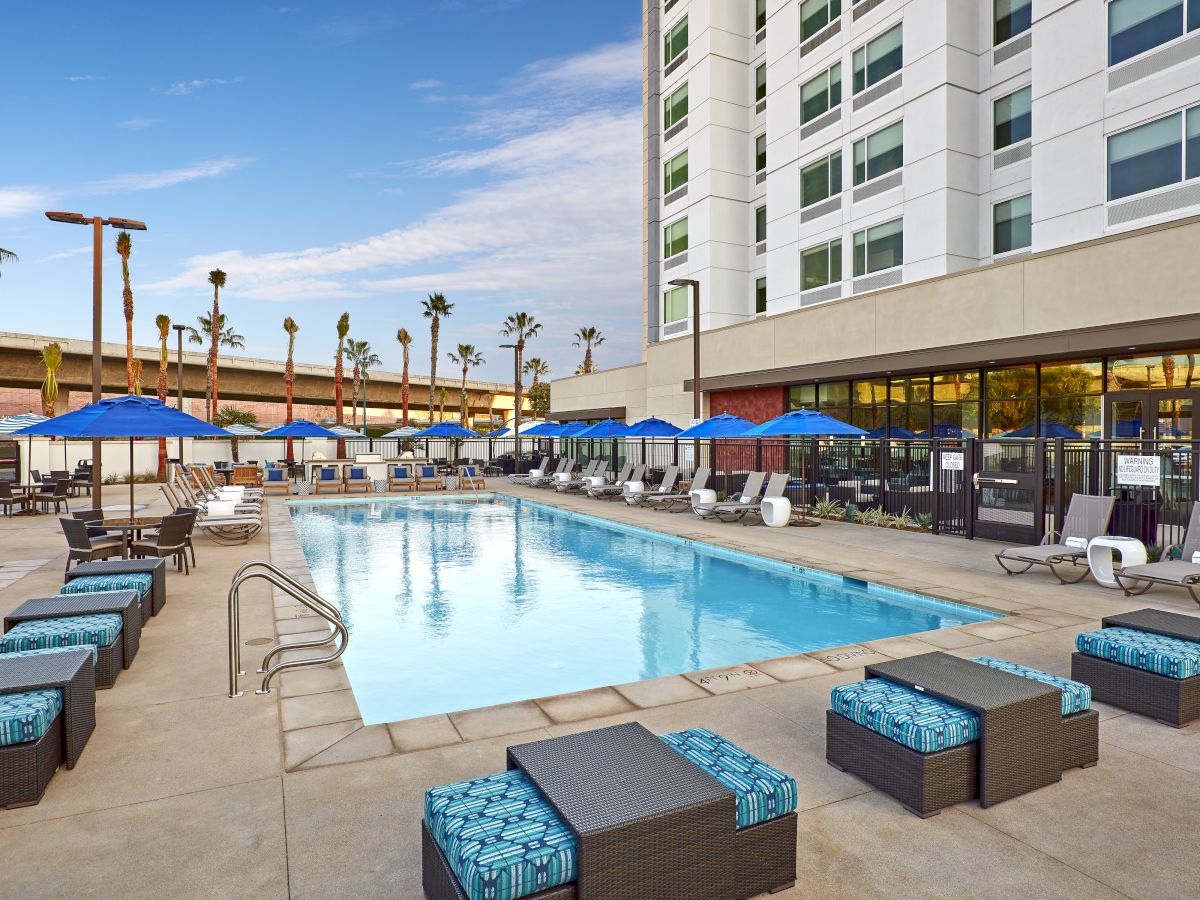 The image shows an outdoor pool area at a hotel with lounge chairs, blue umbrellas, and patio furniture. Palm trees and a clear sky are visible.