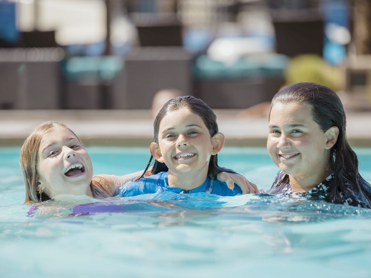 Three children are smiling and enjoying their time in a swimming pool, with their arms around each other.