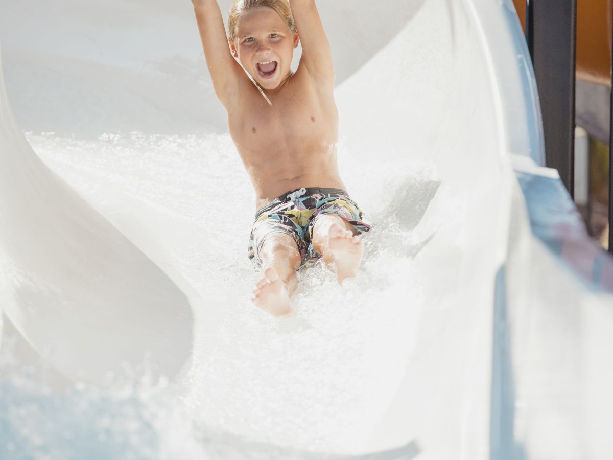 A child is sliding down a water slide, with arms raised and a smile on their face, enjoying the activity in a bright and sunny environment.