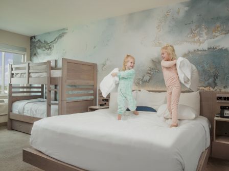 Two young children in pajamas are having a pillow fight on a bed in a room with bunk beds and a large wall mural.