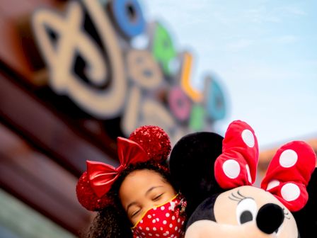 A child wearing a red polka dot mask and Minnie Mouse ears hugging a Minnie Mouse plush, with a blurred Disney-themed background.