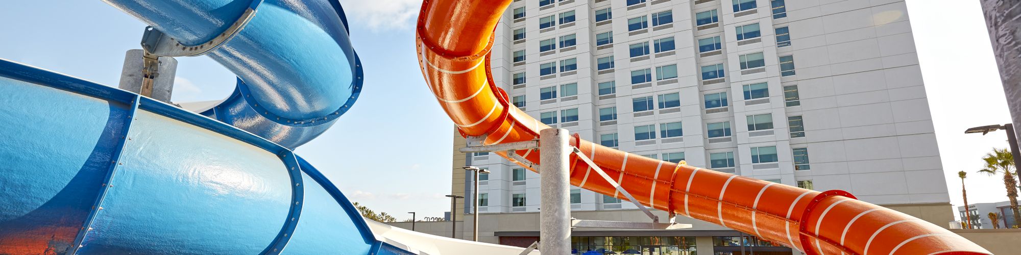 The image features colorful water slides at an outdoor water park with a tall hotel building in the background, under a clear sky.