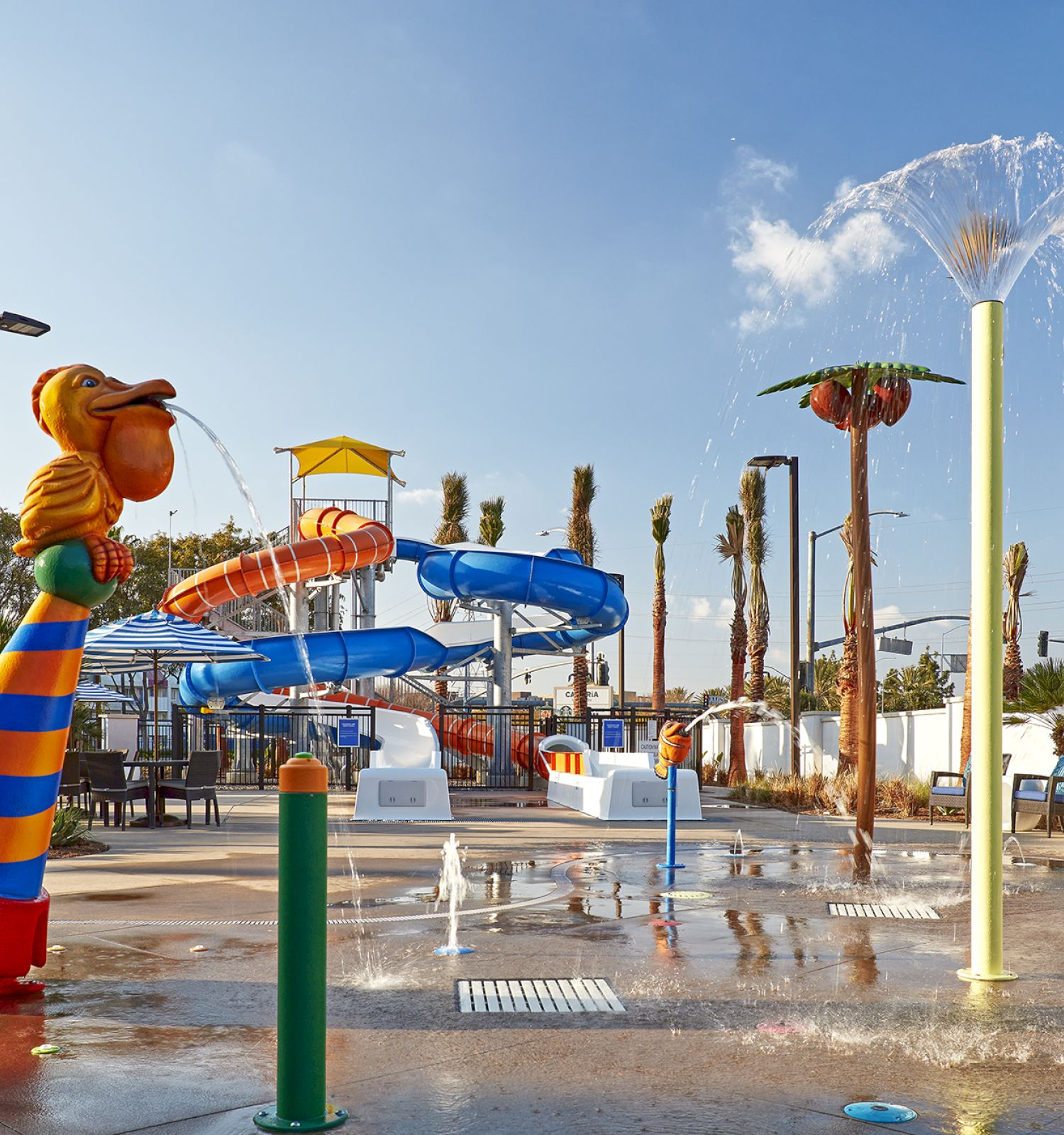 The image shows a colorful water park with slides, spraying fountains, and palm trees under a clear blue sky.