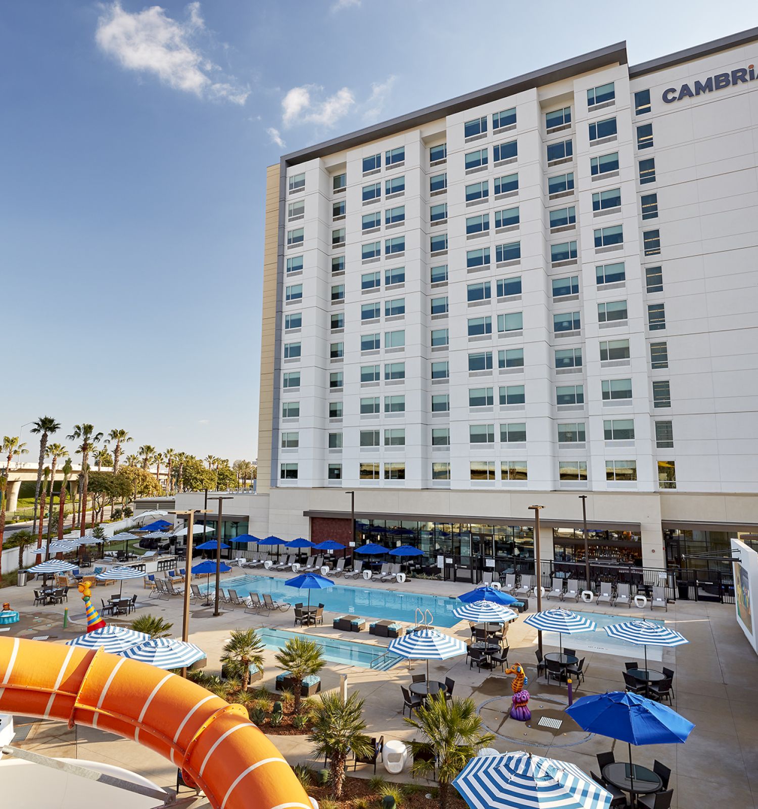 The image shows a tall hotel building labeled "Cambria" with a large pool area, umbrellas, and a water slide in the foreground on a sunny day.