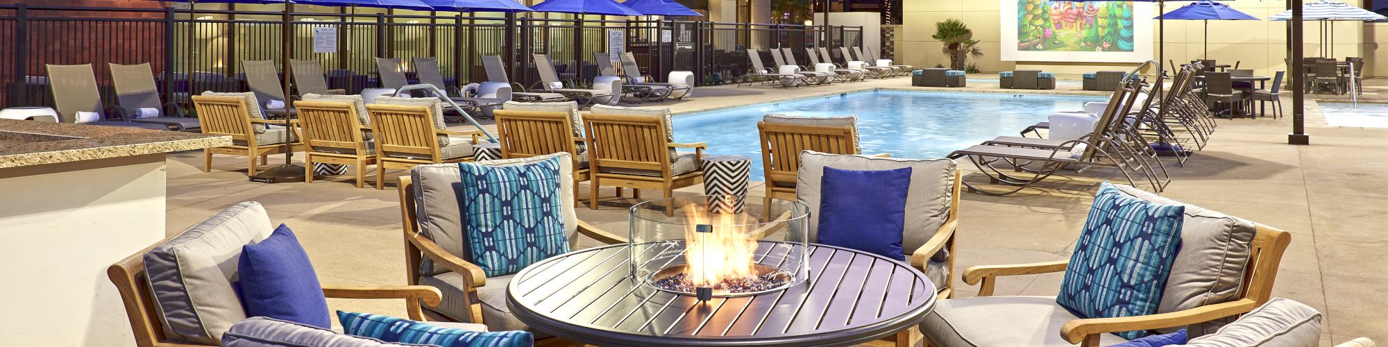 The image shows an outdoor pool area with cushioned wooden chairs, a round fire pit, lounge chairs, and blue umbrellas at a hotel during the evening.