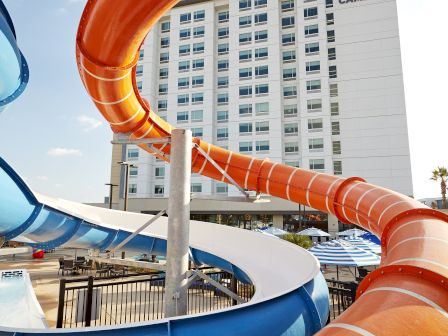 A hotel building stands behind a colorful water slide with orange and blue tubes, set under a bright sky with some clouds.