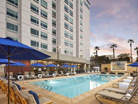 A hotel pool area with lounge chairs, blue umbrellas, and a white multi-story building in the background at sunset. The scene is peaceful and inviting.