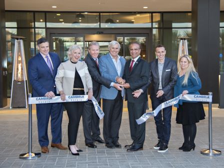 A group of seven people are cutting a ribbon in front of a building entrance, symbolizing the opening of a Cambria Hotel.