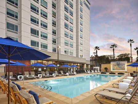 A hotel pool area with blue umbrellas, sun loungers, and a projector screen is adjacent to a multi-story building at sunset with palm trees in the background.