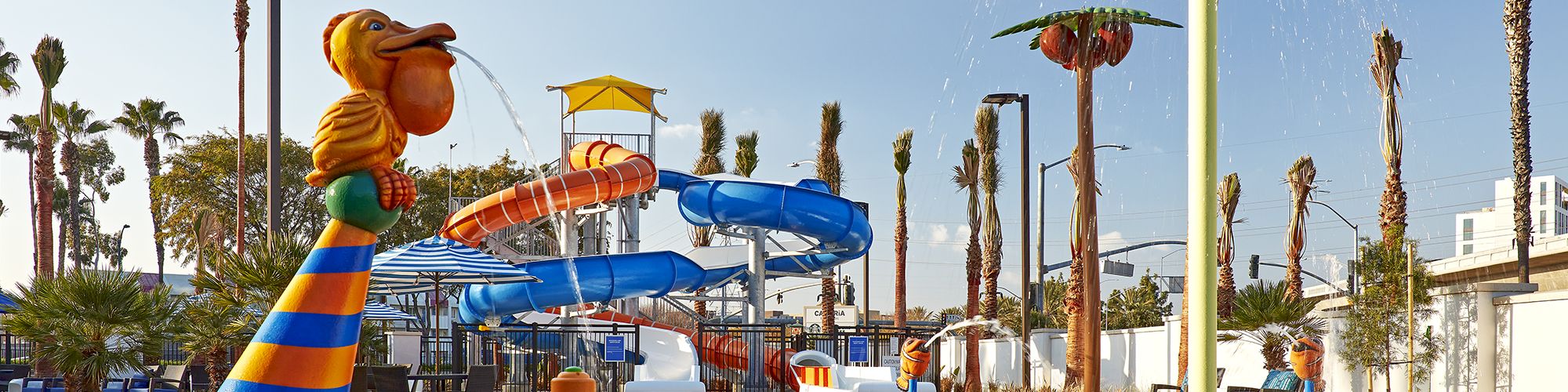 A colorful water playground with slides, a pelican sculpture, and various sprinklers on a sunny day. Palm trees are in the background.