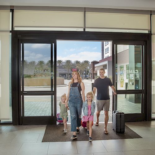 A family with two children is entering a building through glass doors. The adults are carrying bags and one is pulling a suitcase on wheels.