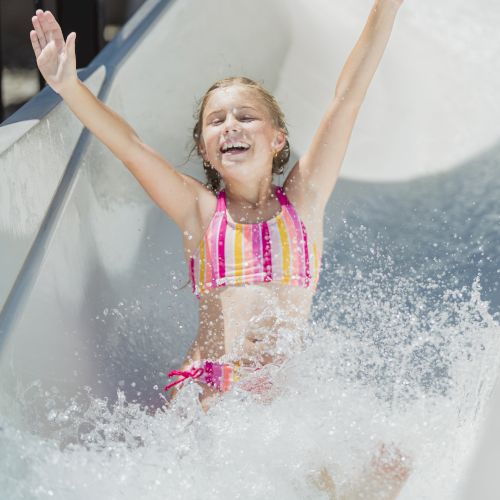 A child enjoying a water slide, arms raised and smiling, wearing a colorful swimsuit, splashing in the water at a park.