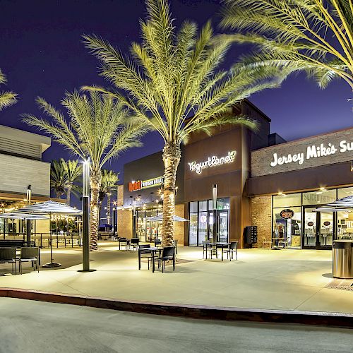 The image shows a shopping center plaza at night with palm trees, outdoor seating, Yogurtland, and Jersey Mike's Subs.