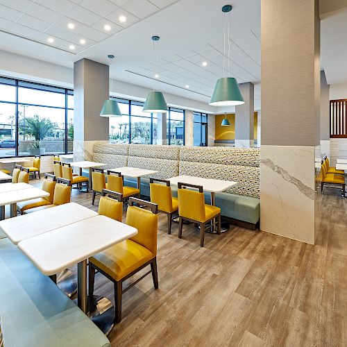 A modern, well-lit restaurant with yellow and green chairs, white tables, and large windows, featuring modern pendant lighting and wood flooring.