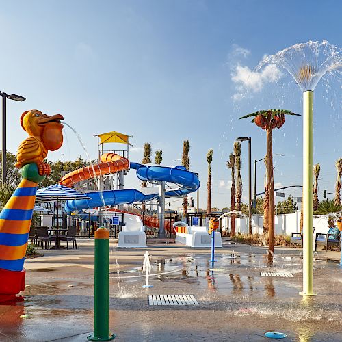 A colorful water park with slides, water features, and sprinklers surrounded by palm trees under a clear sky.