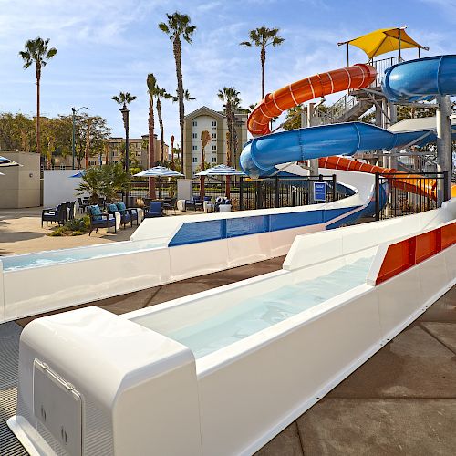 A colorful water slide complex with twists and turns, splash pools, palm trees, and a seating area with umbrellas.