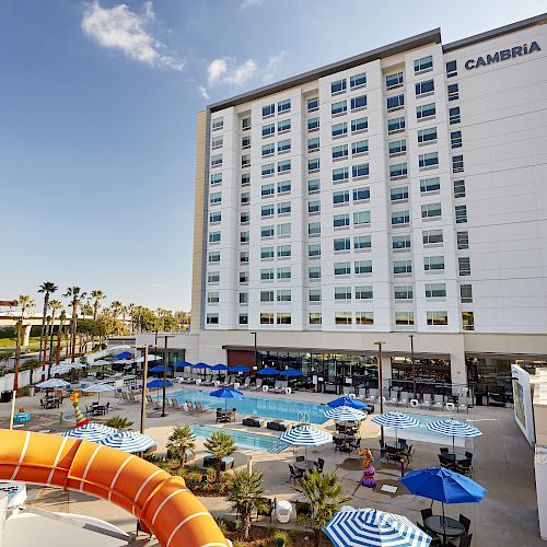 A tall hotel building with "Cambria" branding, featuring an outdoor pool area with umbrellas, lounge chairs, and a waterslide in the foreground.