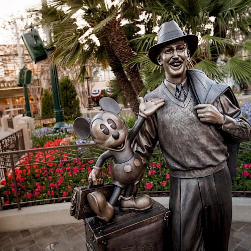 A statue of a man with a hat and suitcase standing next to a Mickey Mouse statue in a garden area with flowers and trees, in what looks like a park.