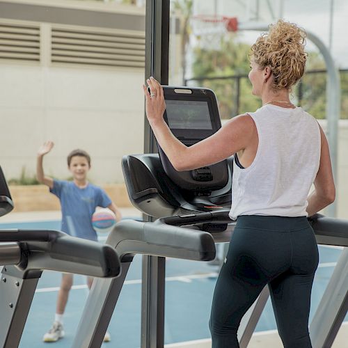 A woman on a treadmill inside a gym waves to a child playing basketball outside the window.