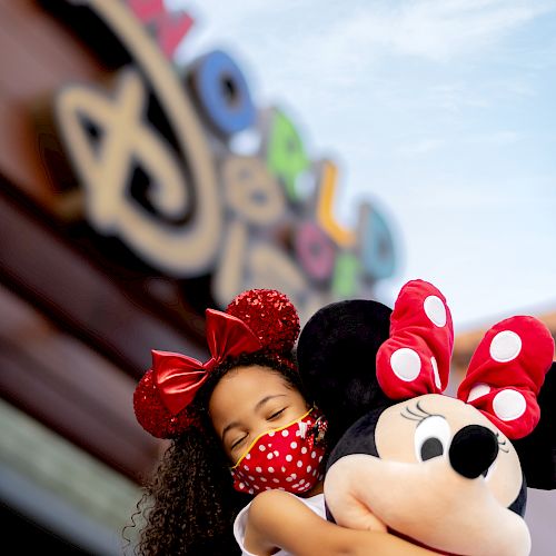 A child wearing a red bow and face mask hugs a Minnie Mouse toy in front of a colorful sign, possibly at an amusement park.