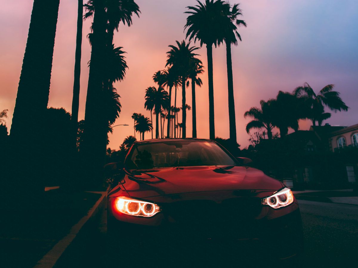 A car with its headlights on is parked on a street lined with palm trees, with a colorful sky at sunset in the background.