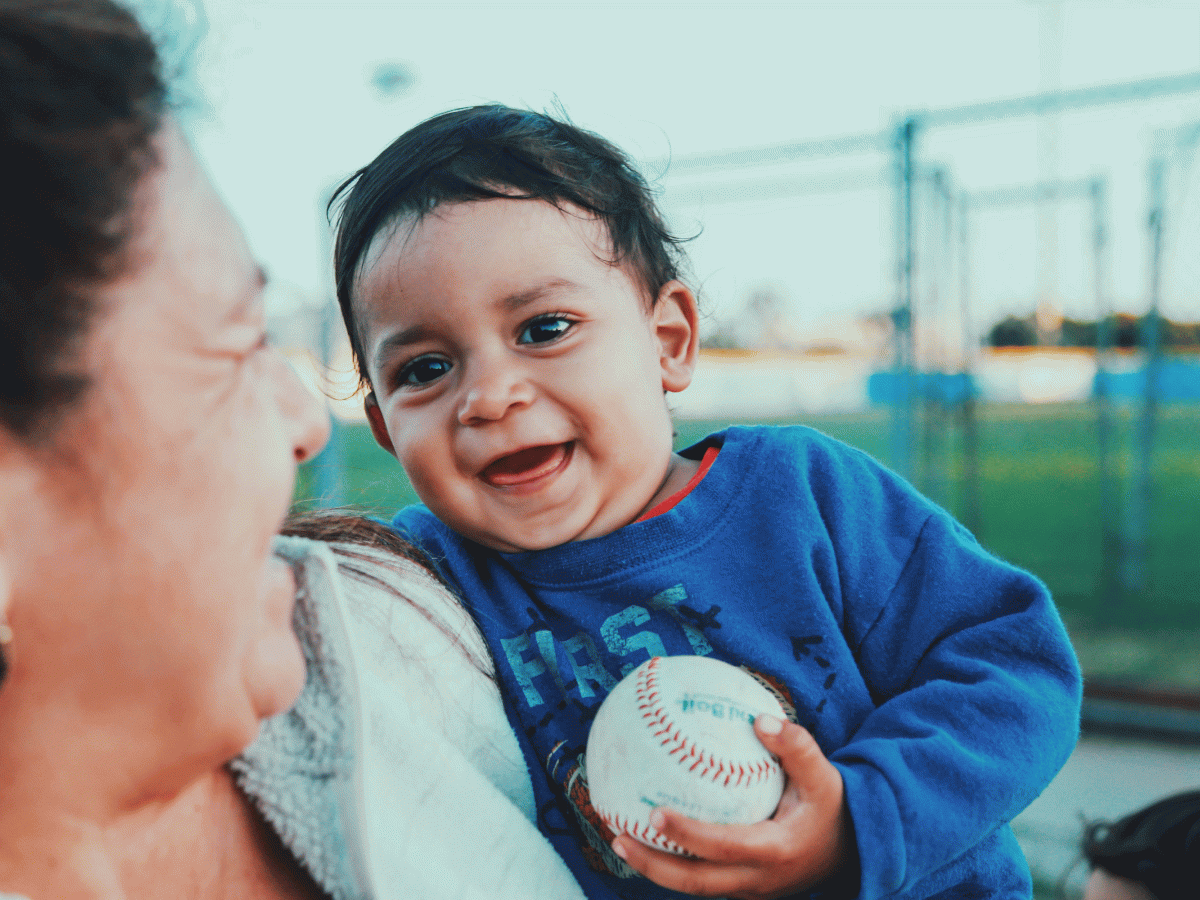 A smiling child holding a baseball while being held by an adult outside near a field, both enjoying the moment at what appears to be a sports venue.