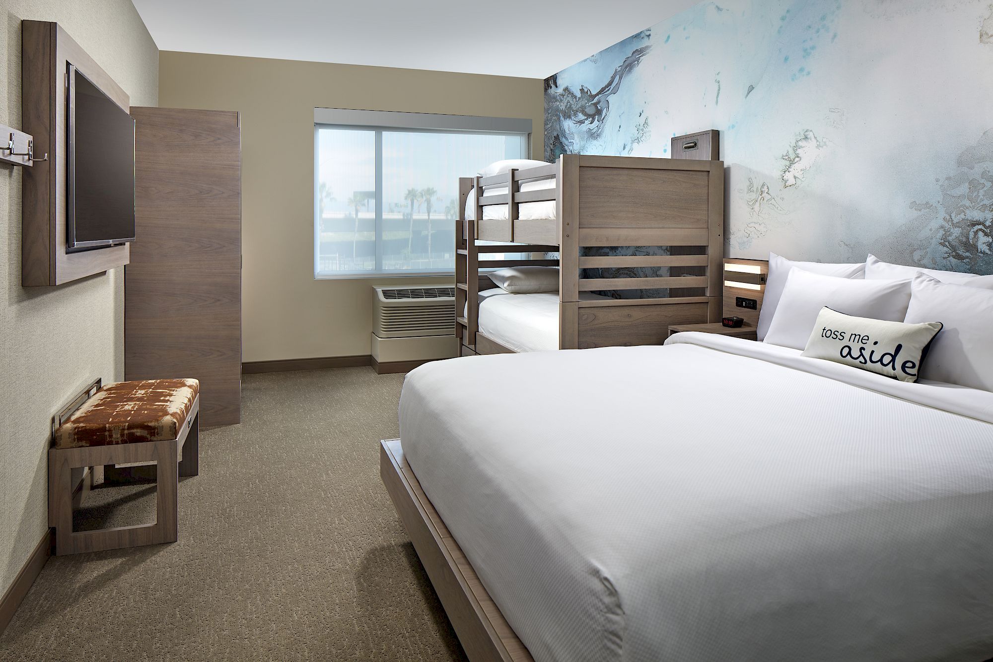 The image shows a clean, modern hotel room with a queen bed, a bunk bed, a flat-screen TV, a bench, and a window with blinds, all on a carpeted floor.