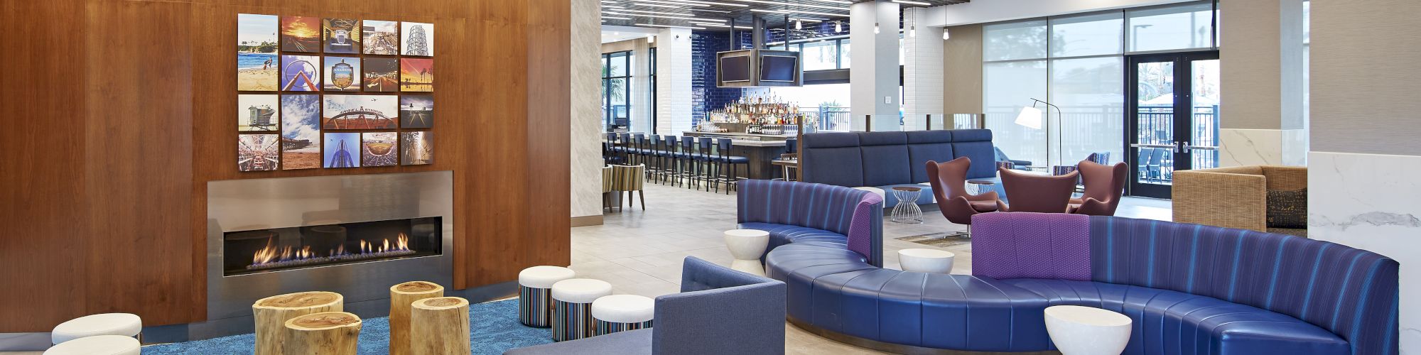 The image shows a modern lobby with seating, a fireplace, blue chairs, and wooden accents. There is ample lighting and a bar area in the back.