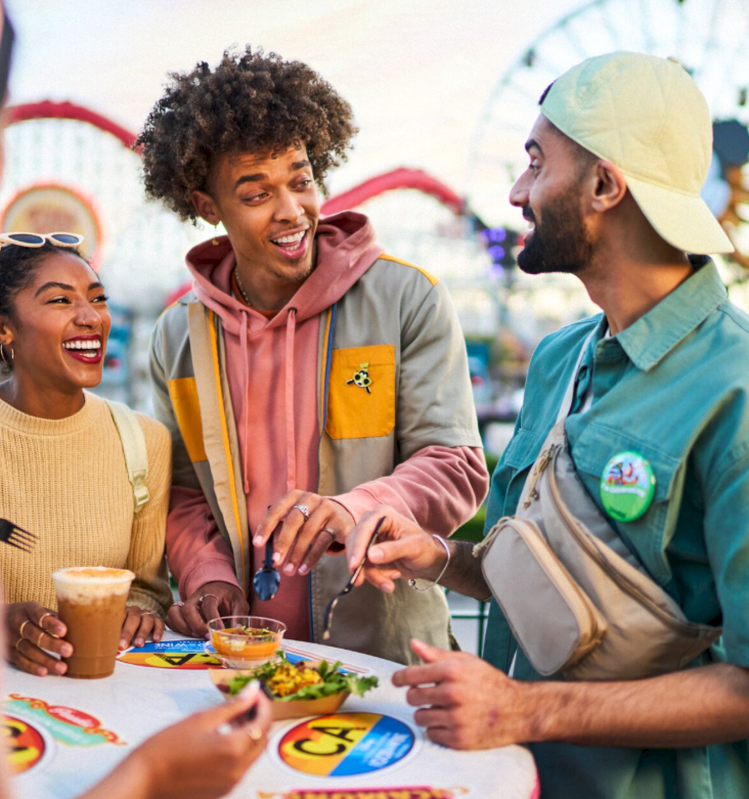 A group of friends enjoying food and drinks, laughing together outdoors at what appears to be an amusement park.