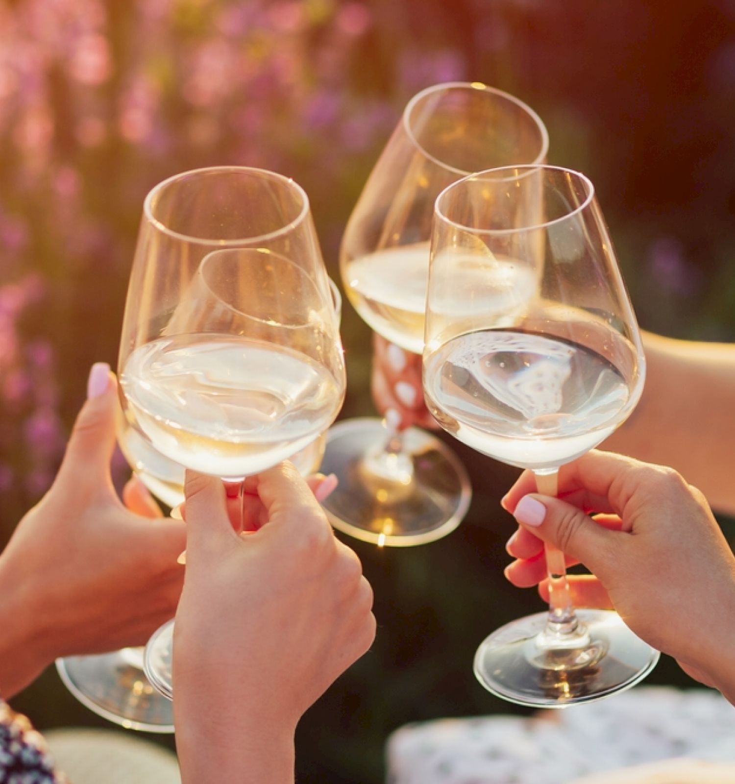 Three people clink glasses of white wine together outdoors, with a background of blurred, sunlit flowers.